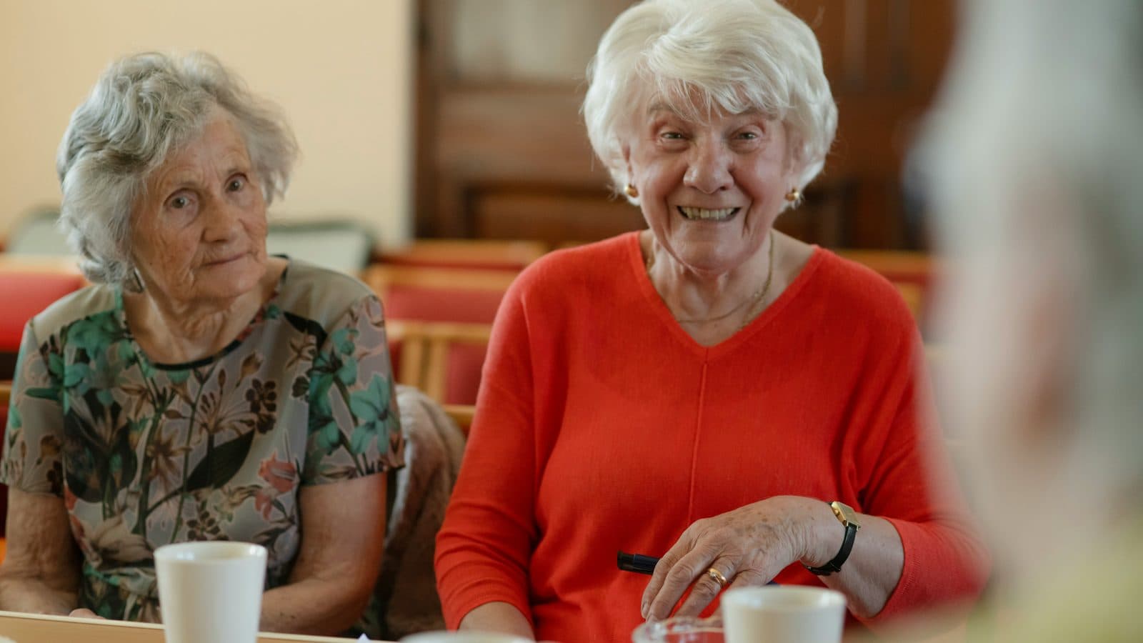 Two older women seated at a table with cups of tea and a sheet of paper, one smiling at someone across the table.