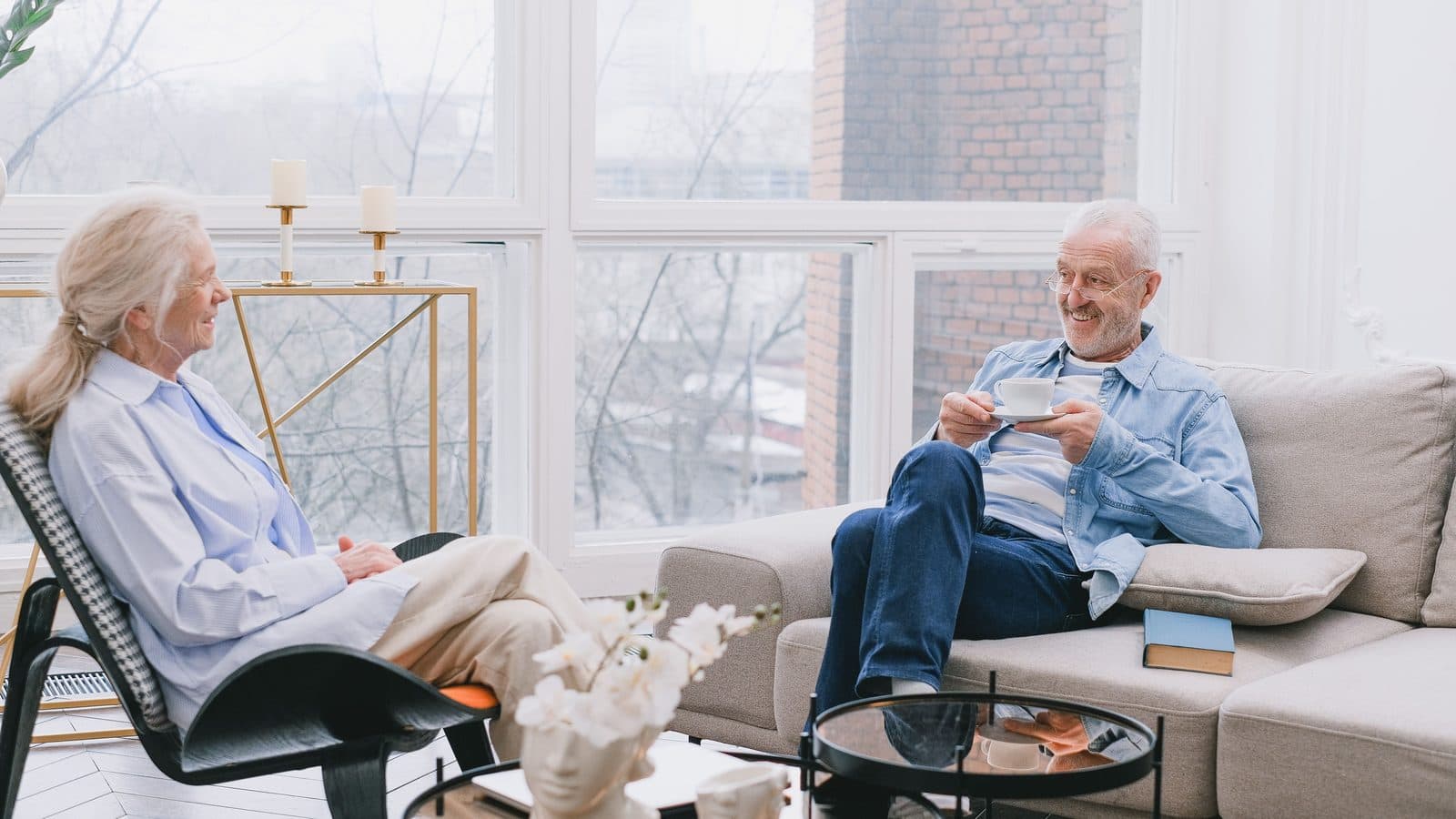 An older couple sitting and talking in a bright, plainly furnished common room.