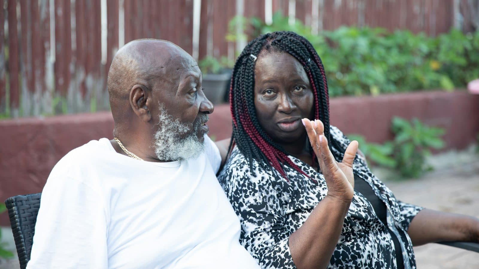 An older man and woman sitting together in a garden, mid-conversation, in front of a red wooden fence.
