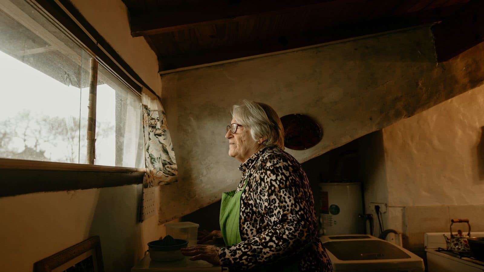 An older woman standing at the window of her own home, looking out.