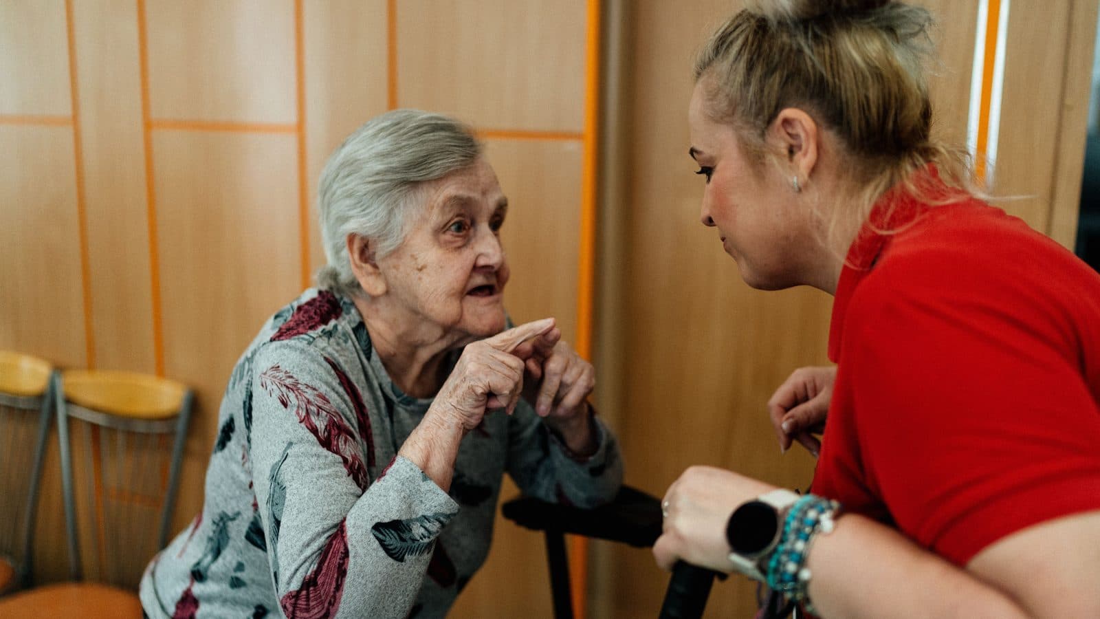 A younger woman leaning in to talk with an older woman seated at home.