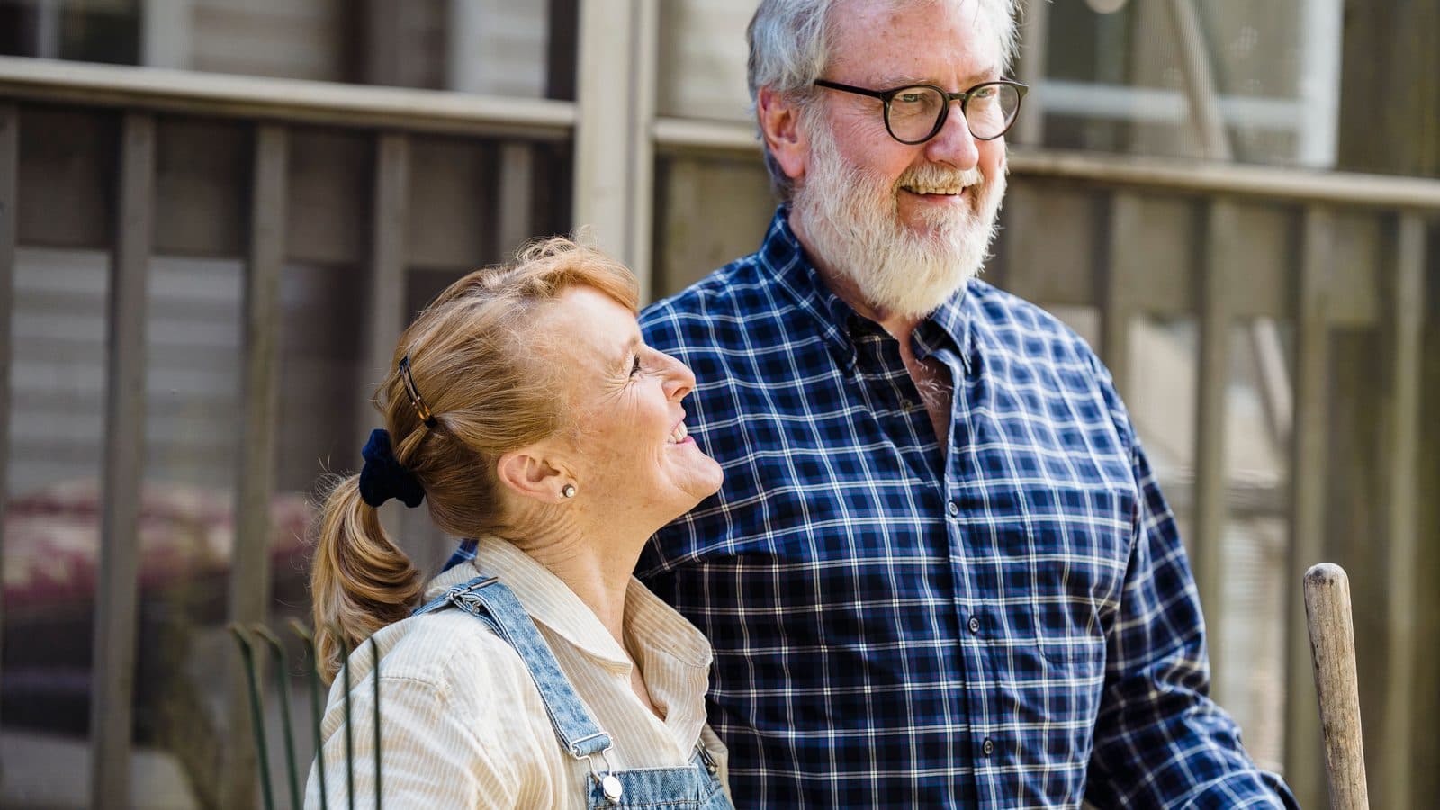An older couple standing outside their home with garden tools, looking at each other.